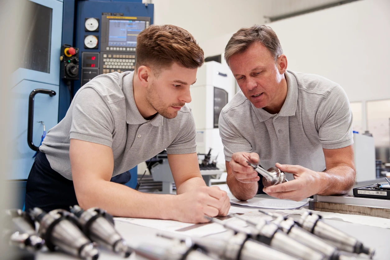 Two men in grey shirts looking down at a small metal component which one of them holds.