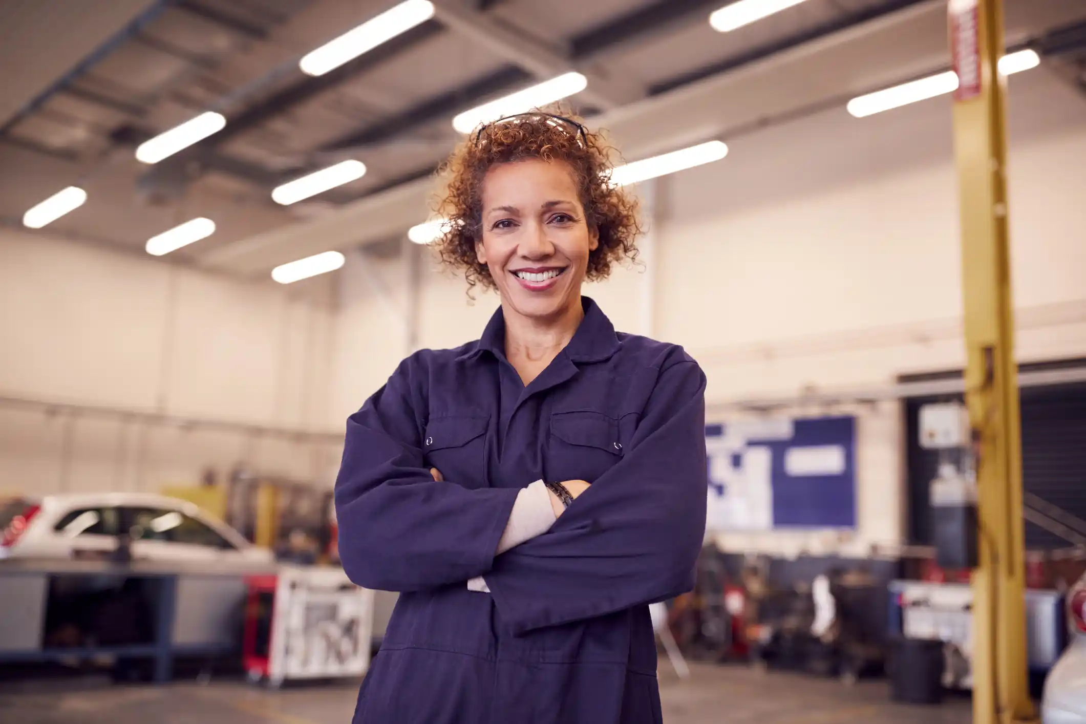 A person in blue overalls standing in a garage smiling with arms folded.