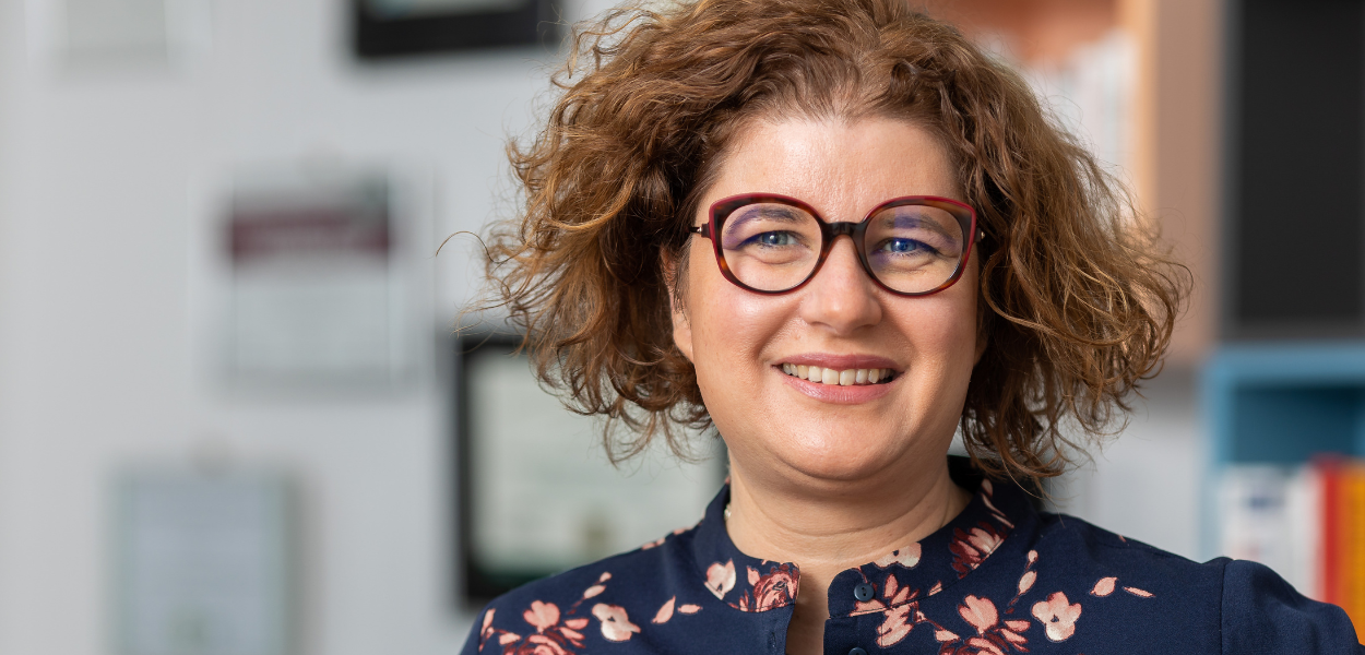 Photo of a woman, the CISO. She looks into the camera and smiles. She has curly hair and wears glasses.