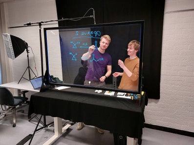 Thomas Hansen and Jaap Harteveld draw on the lightboard (a transparent glass board) while standing behind it in front of a black backdrop