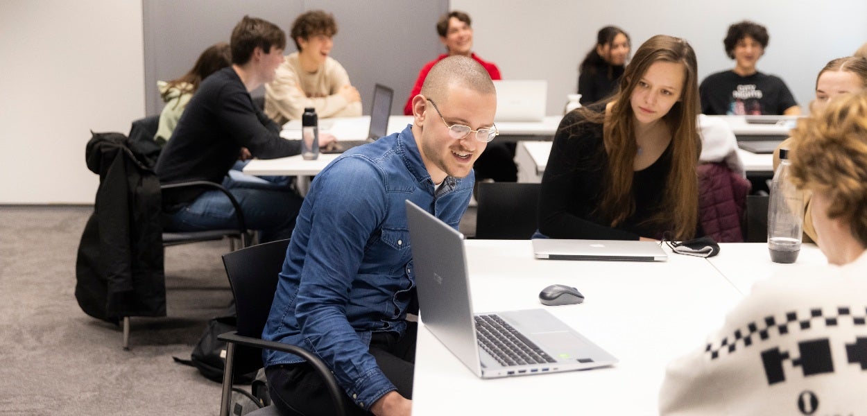 Een ruimte vol studenten met op de voorgrond een groepje van drie meiden en een man die kijken naar een laptopscherm.