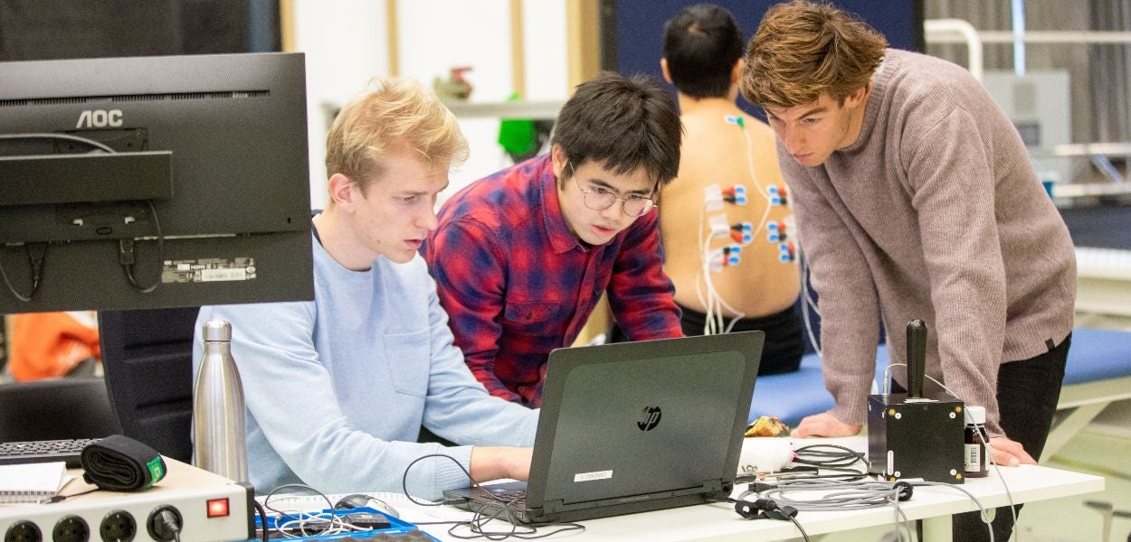 Drie mensen werken samen aan een laptop, met op de achtergrond iemand met medische sensoren op zijn rug.