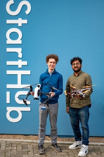 Aleksey Martynyuk en Aryan Sharma poseren met hun 3D-geprinte drones in de hand voor de hemelsblauwe buitenmuur van de VU StartHub