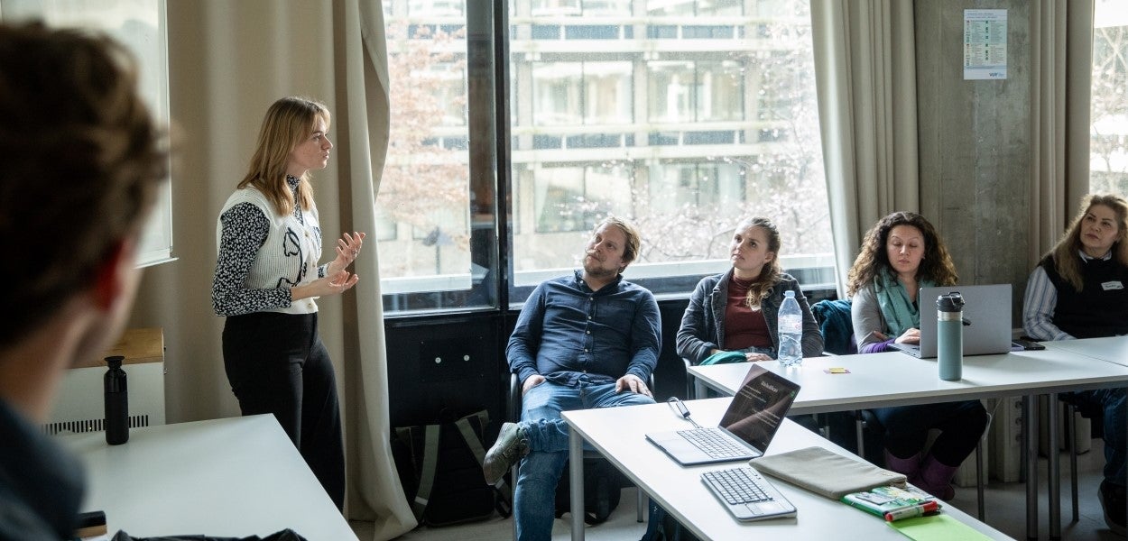 Een vrouw staat voor een klein groepje aandachtig luisterende mensen