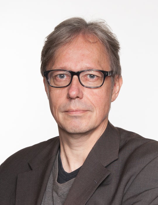 Portretfoto van een man met grijs haar en donkere bril, gekleed in een donker beige jasje en grijs shirt
