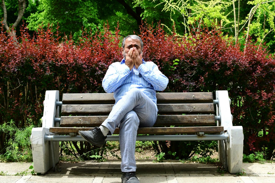 A man sits on a bench outside and sneezes.