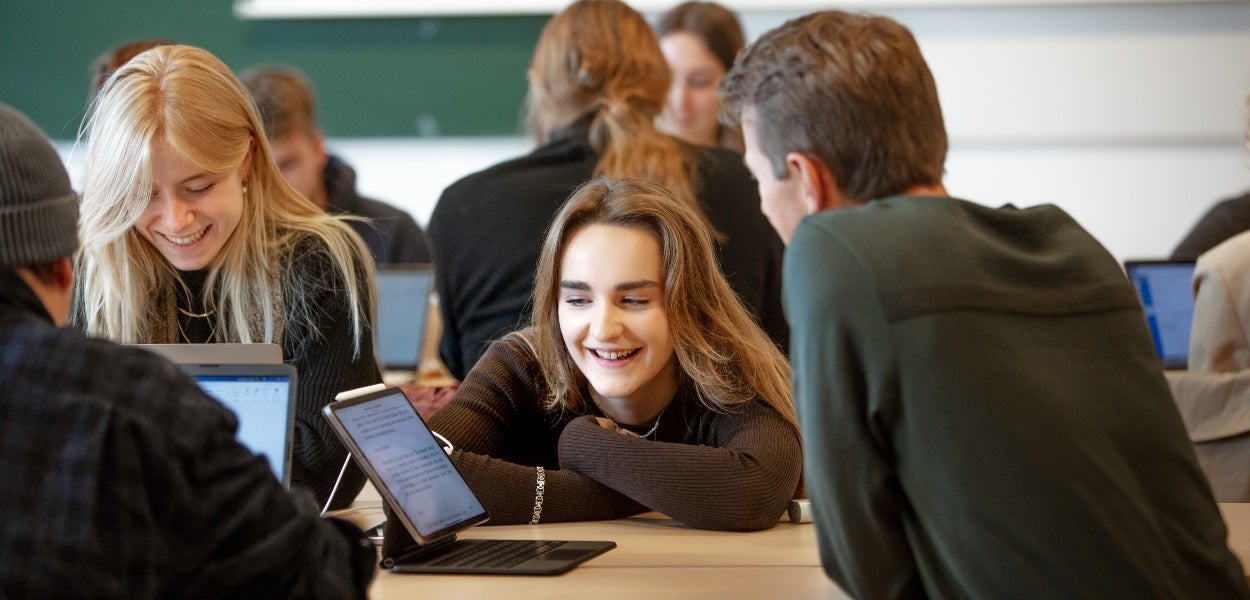 In een volle ruimte kijkt een vrouw met blond haar lachend richting het scherm van een tablet.