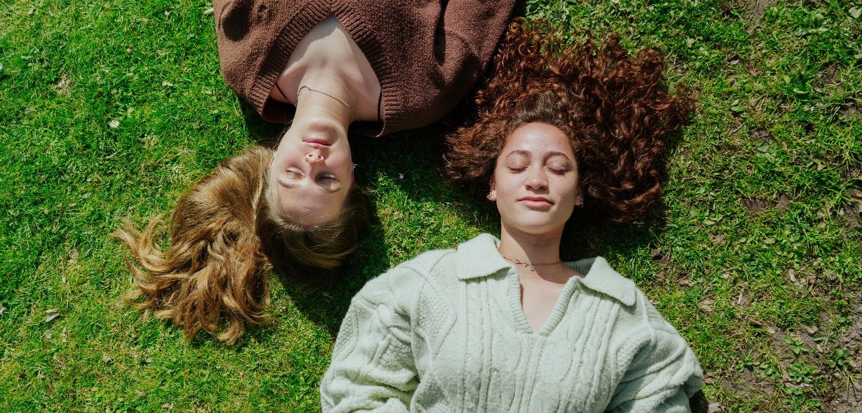 Twee vrouwen liggen rug aan rug op gras, met hun ogen gesloten, genietend van de buitenlucht.