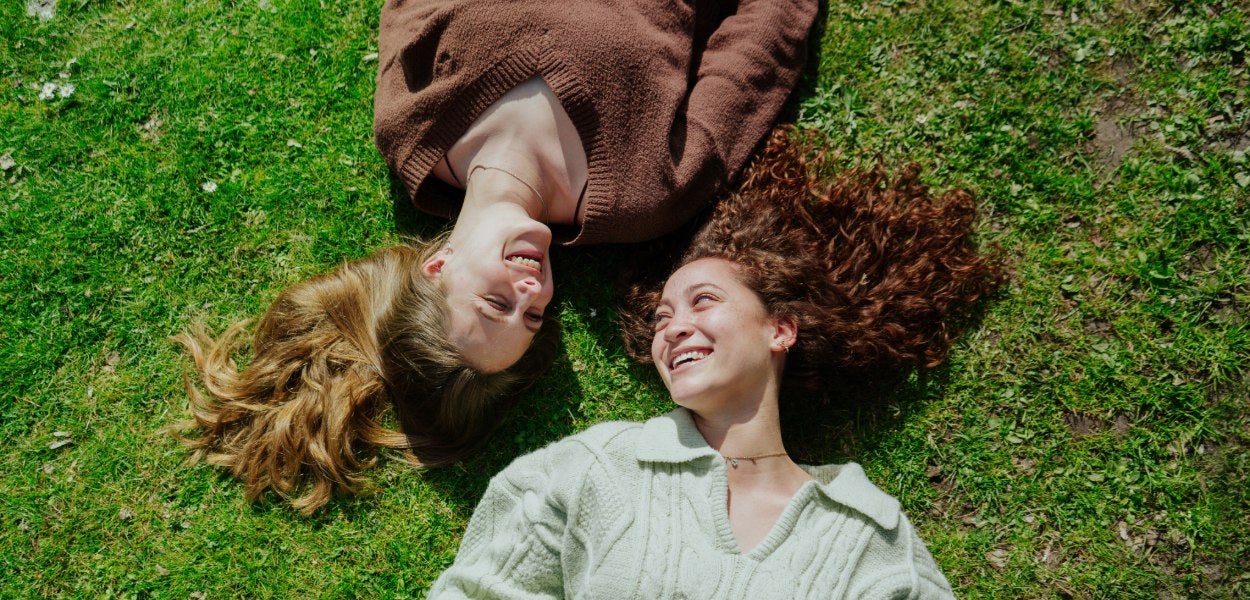 Two women lie side by side on the grass, while smiling to each other.