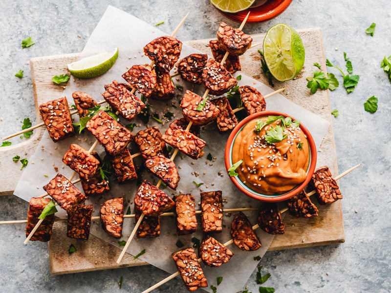 Plank met tempeh aan satéprikkers en een bakje met een oranje saus.