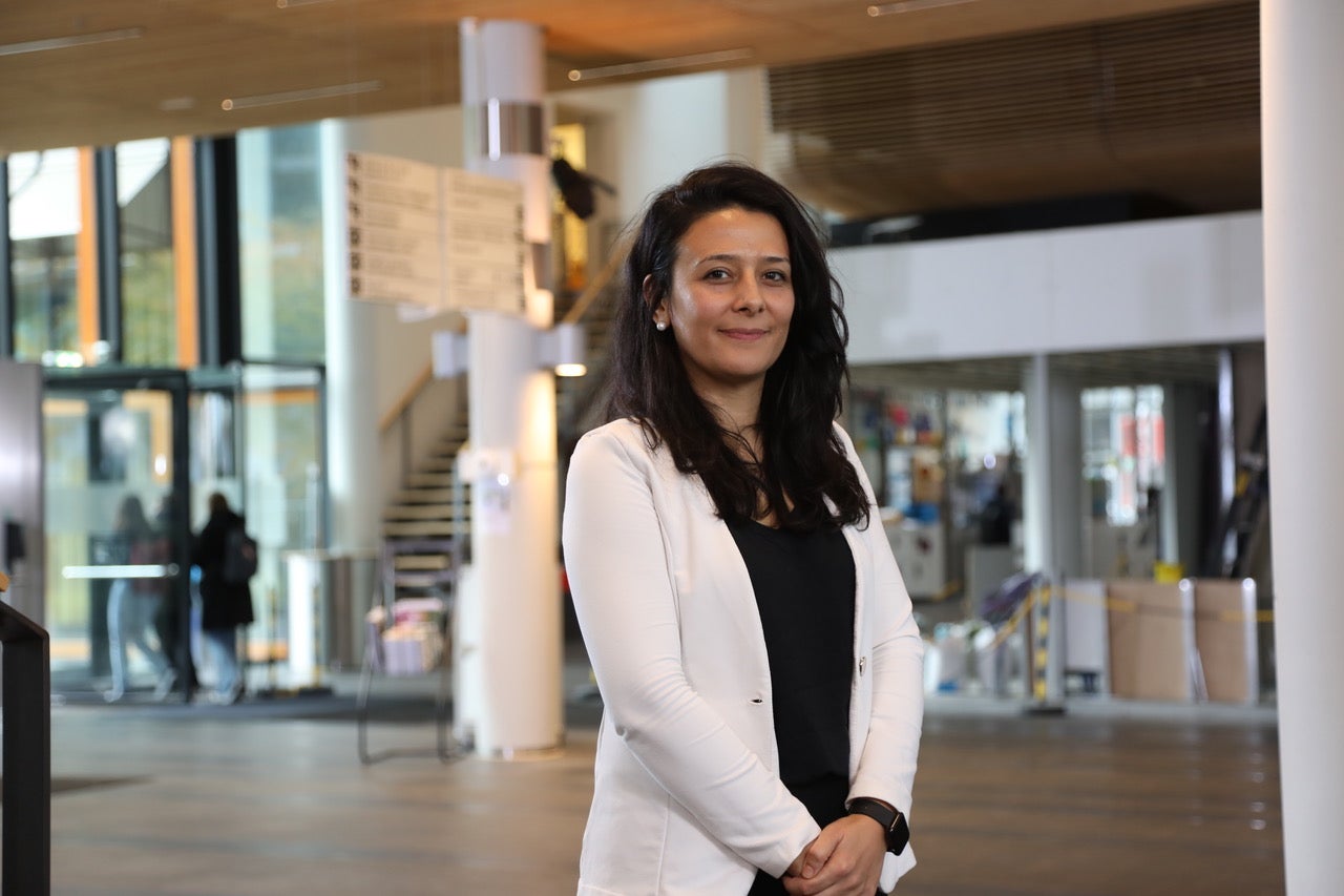 Smiling woman with black long hair dressed in white jacket over a black shirt stands near a building or courtyard