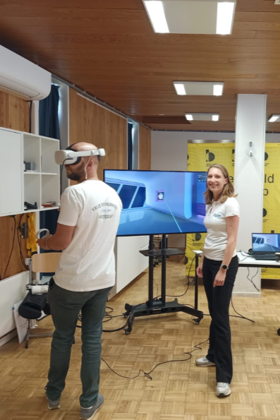 Een man met een grijze spijkerbroek kijkt door een VR-bril, terwijl een blonde vrouw lachend de camera inkijkt. Achter hen bevinden zich diverse banners en een scherm dat de VR-omgeving weergeeft.