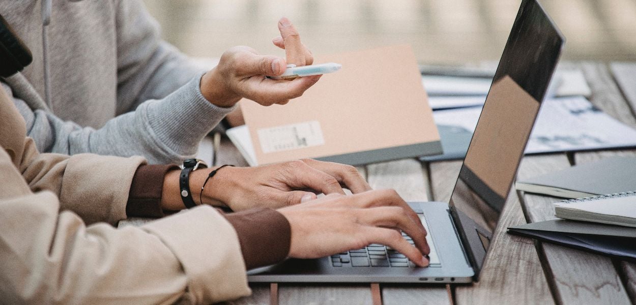 Two people working together on a laptop, with one person typing and the other pointing at something with a pen.