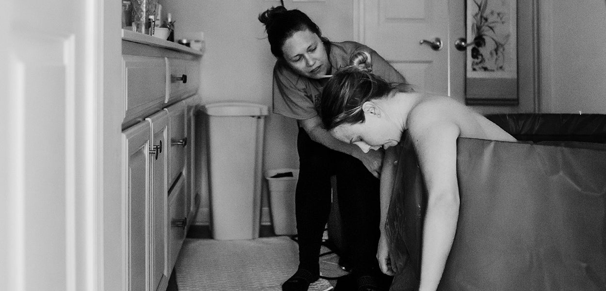 Zwartwit foto van een vrouw in bad, ze leunt voorover, tijdens een bevalling. Een midwife begeleidt haar.
Foto door Rebekah Vos