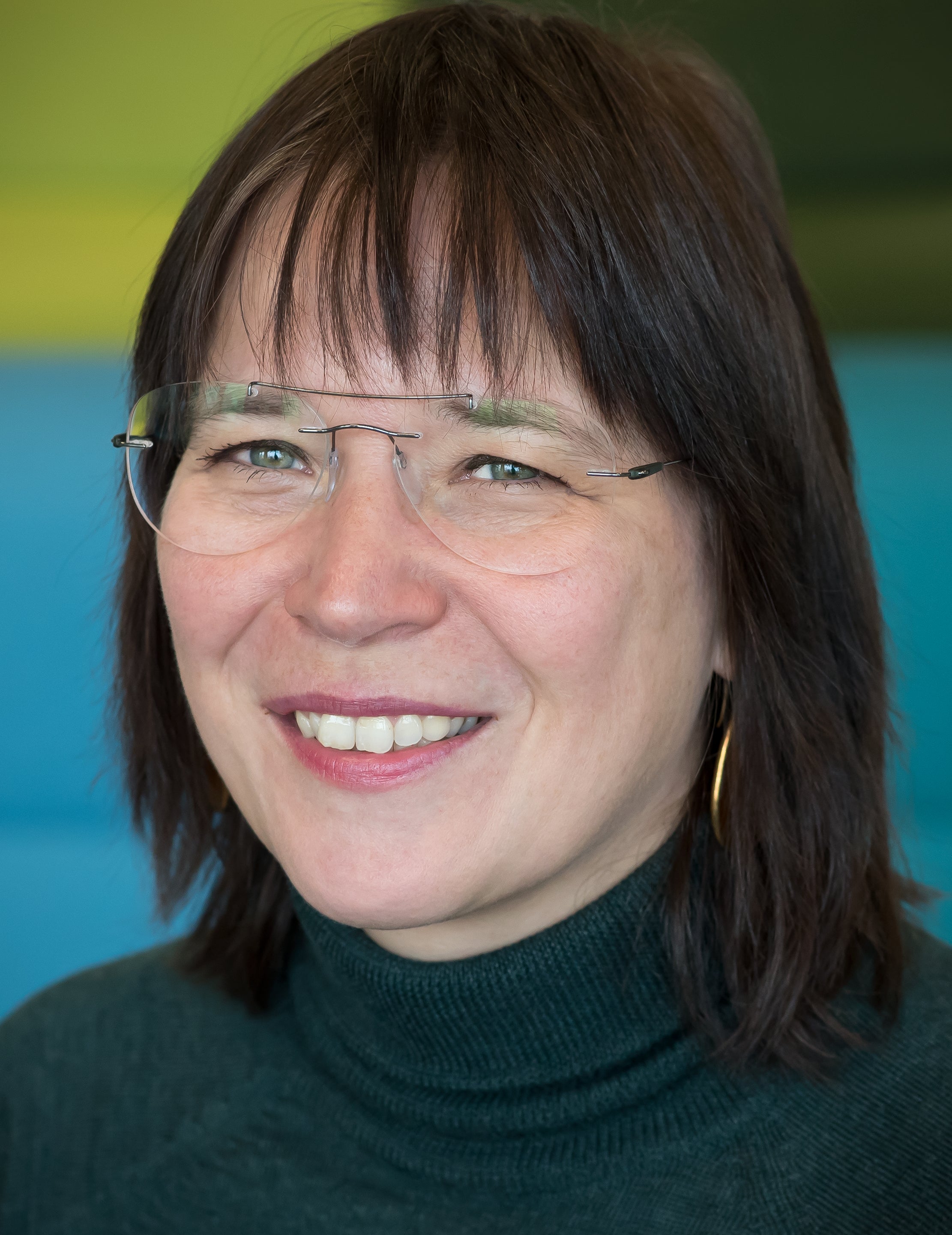 Portrait photograph of smiling woman with dark brown medium length hair glasses and gold earring, dressed in a dark green turtleneck.