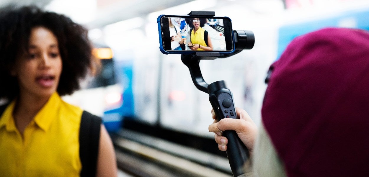 Person filming a woman with a smartphone on a gimbal at a train station.