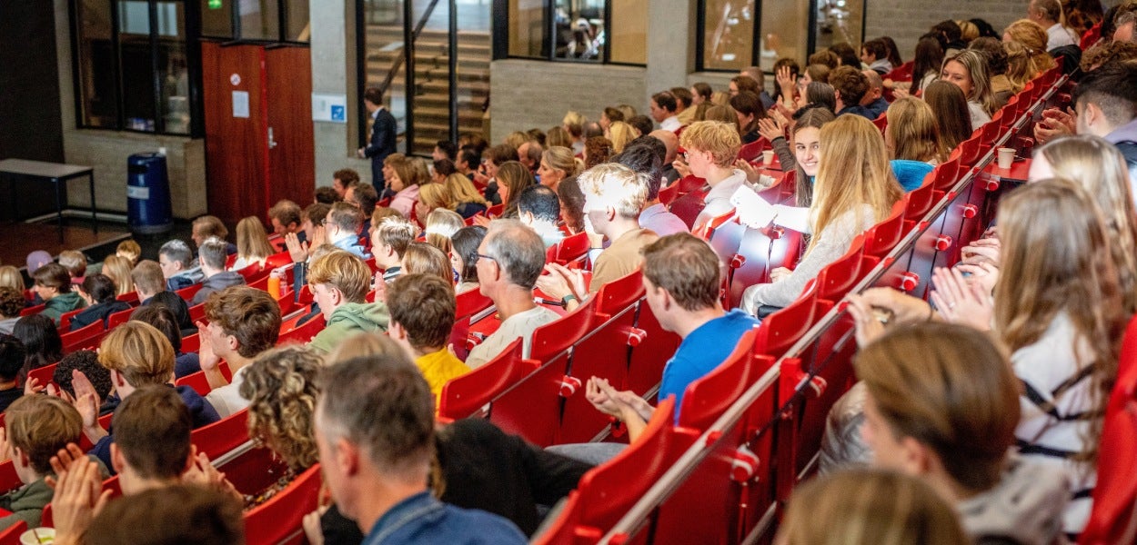 A group of people sit in a lecture hall with red chairs.