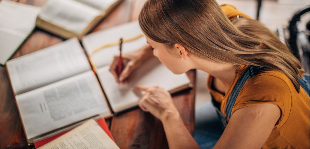 A women with dark blond hair is writing in a notebook on a table filled with study books