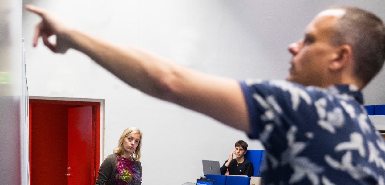 Een man met een blauw-wit shirt aan wijst naar het bord terwijl een vrouw met blond haar en een student achter een laptop toekijken.