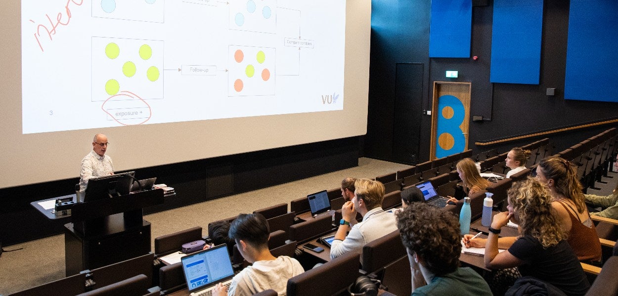 A teacher speaks to students in a lecture hall, with a presentation in the background