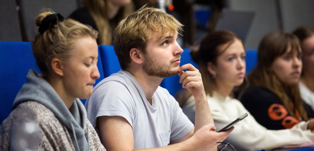 Een jongen met blond haar kijkt voor zich uit, terwijl hij in een collegezaal zit met zijn telefoon in zijn hand.