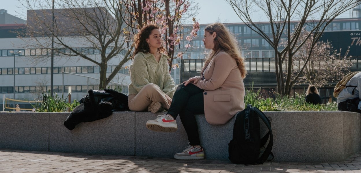 Twee vrouwen zitten op de rand van een grote plantenbak. Omringd door gebouwen en bomen in bloei kijken zij aandachtig naar elkaar.