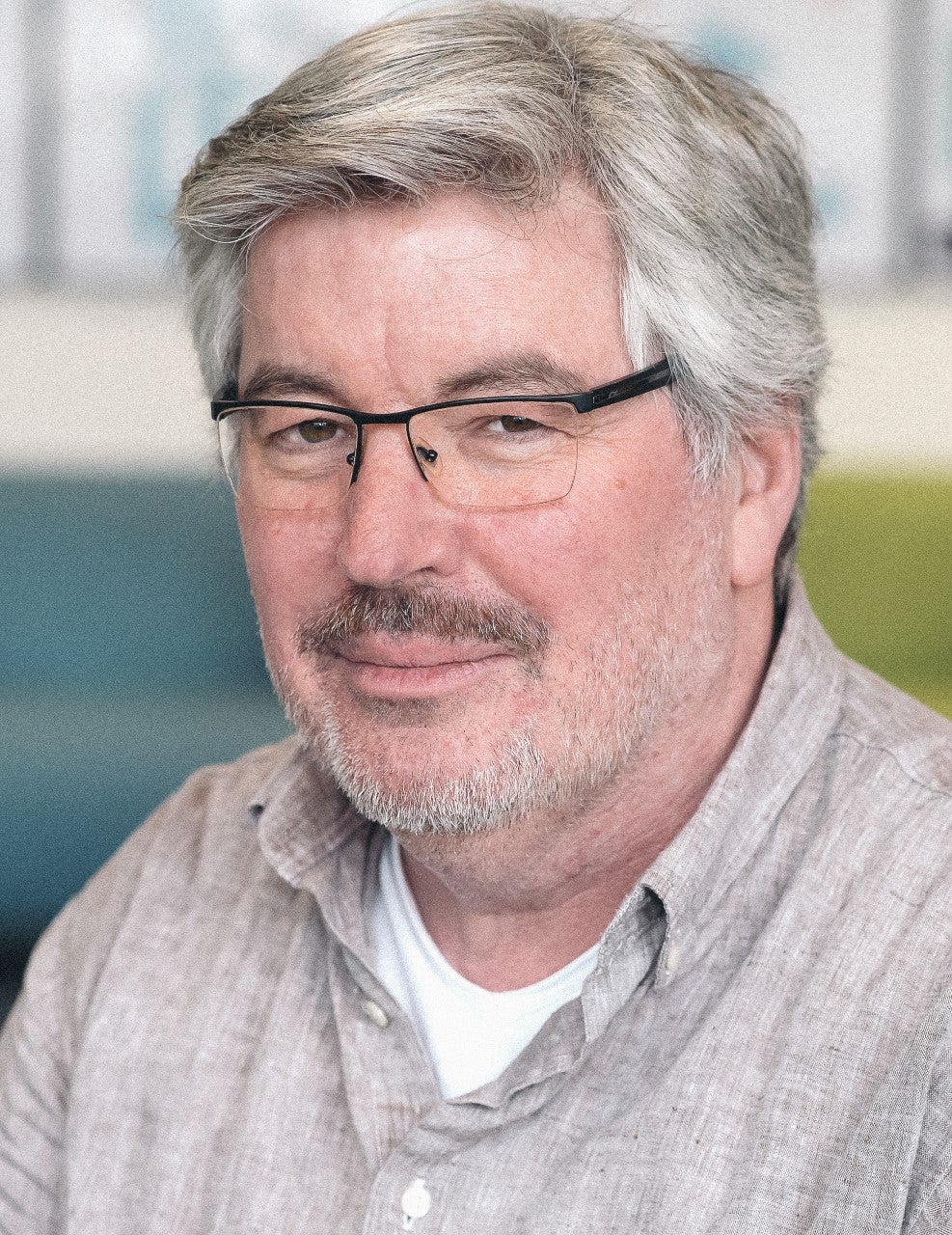 Portret van man met grijs haar en bril met donkere snor en grijze stoppelbaard gekleed in een grijs overhemd en wit t-shirt.
