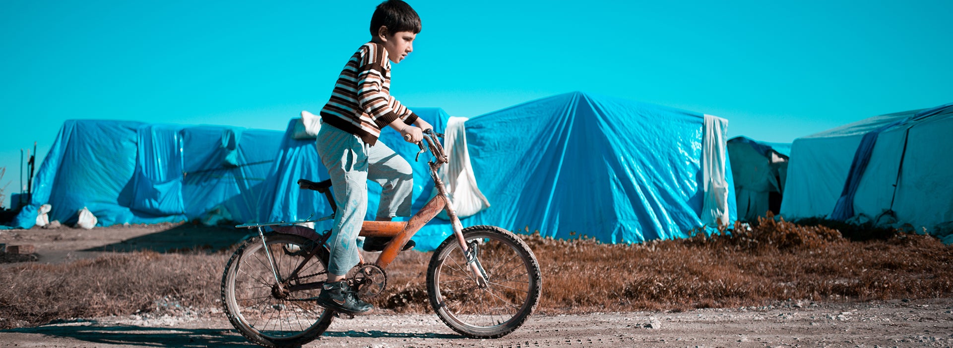 Een jongen fietst langs een vluchtelingenkamp