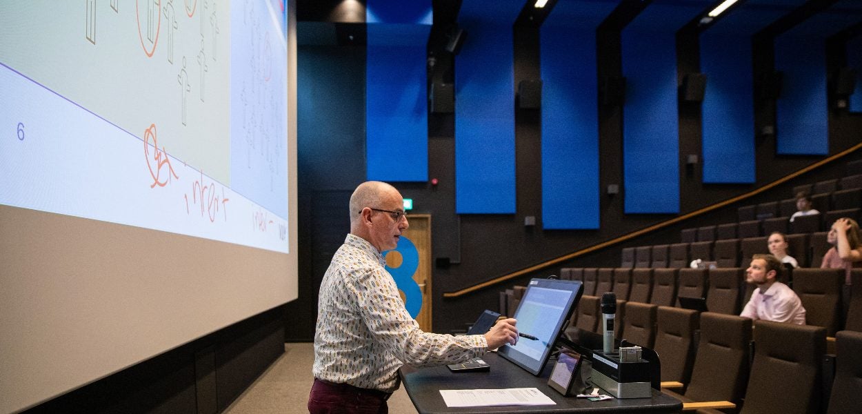 A man is giving a lecture in a modern, mostly empty lecture hall. He stands at a podium in front of a large screen displaying a slide with diagrams and annotations.