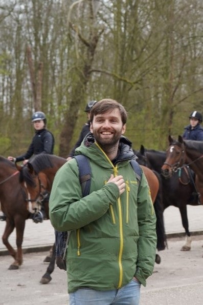 People are casually talking while standing on a path with a group of equestrians in the background.