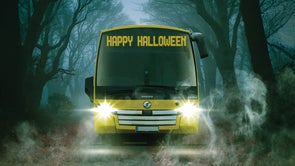 Image is of a bus which says Happy Halloween on its display screen while it drives through a spooky wooded road