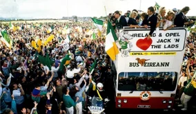 Outdoor shot of a large crowd of Irish Football supporters, cheering and waving flags welcoming the Irish Team home from Italia 90 who are aboard a Bus Éireann open top bus.

The words "IRELAND & OPEL JACK 'N' THE LADS", is prominently displayed in the center of the bus which is adorned with Irish flags and other patriotic symbols. 