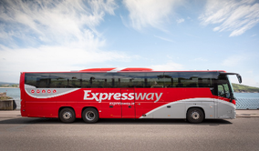 The image shows a long red and white Expressway coach bus parked with the sea in the background