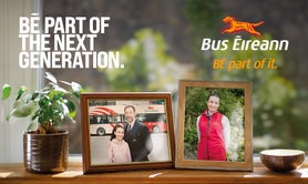 The image is a part of a recruitment campaign for Bus Éireann. The central focus is on a windowsill where two framed photographs are prominently displayed. The first photo shows a man in a suit and a young girl, standing in front of a Bus Éireann bus. The man is the grandfather of the girl; he has his arm around her. The second photo shows the same girl from the first photo, but now older, wearing a red Bus Éireann uniform jacket. 