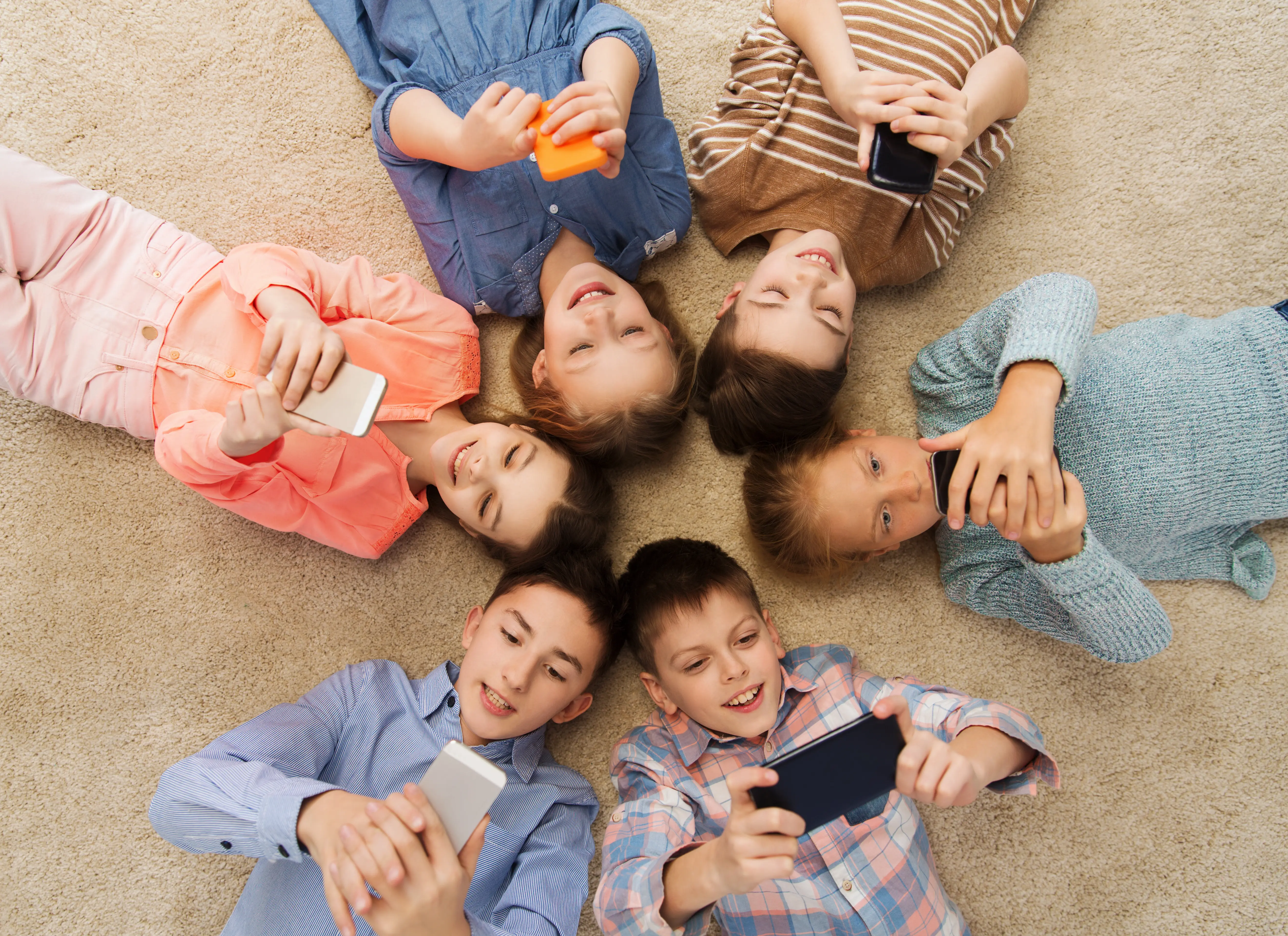 En la imagen, un grupo de seis niños se encuentran en el suelo del living de su casa jugando en su celular. Representación de cómo a la generación alpha tiene interés por los juegos colaborativos y tener experiencias físicas en conjunto a lo digital.