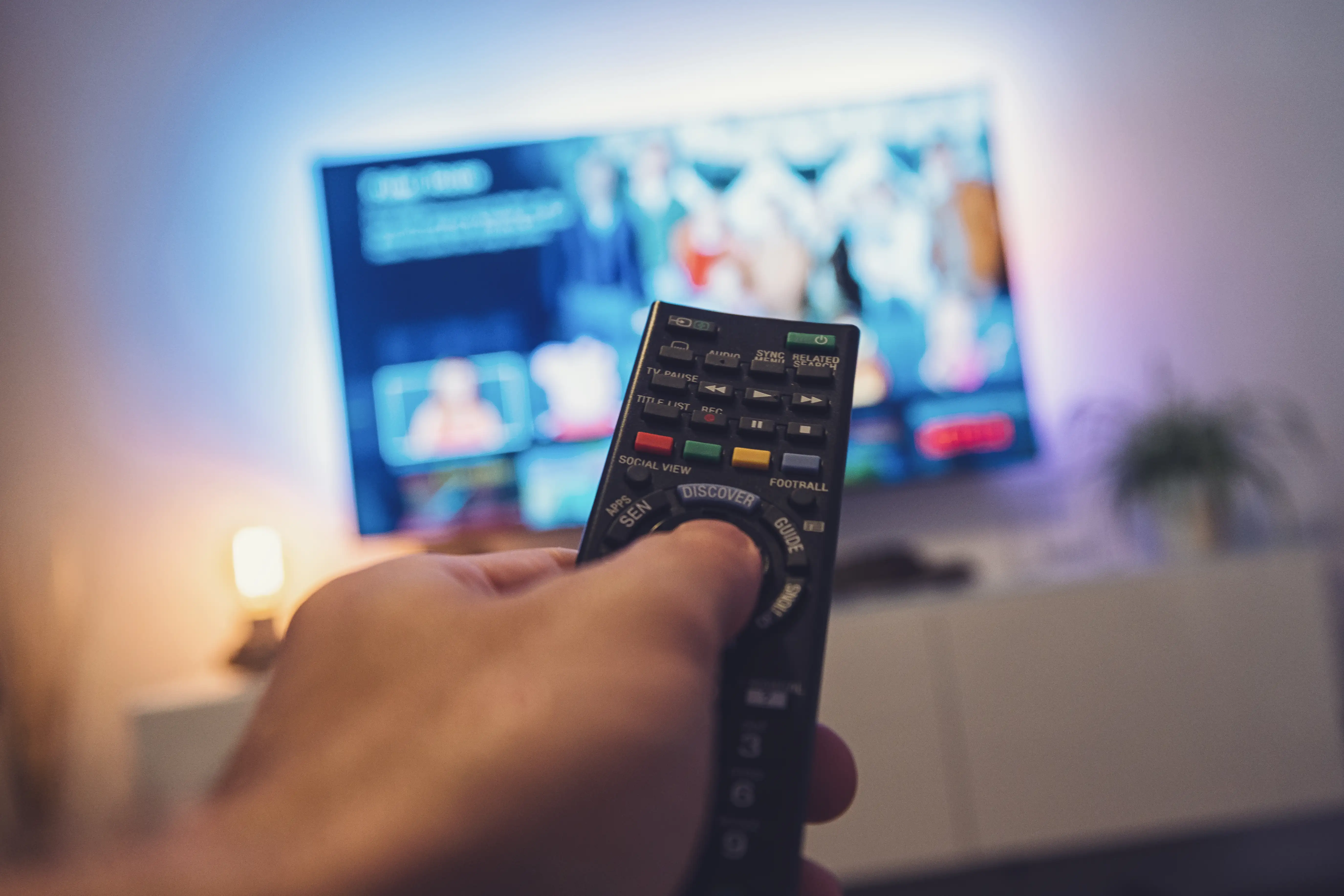 En la fotografía, un hombre sosteniendo un control remoto apunta a la TV donde se reproducen videos en plataforma de streaming.
