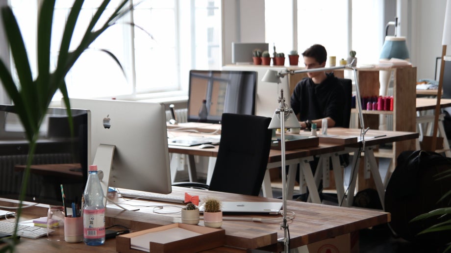 In an office, an individual is sitting down and working at their desk with an Apple Mac computer in front of them.