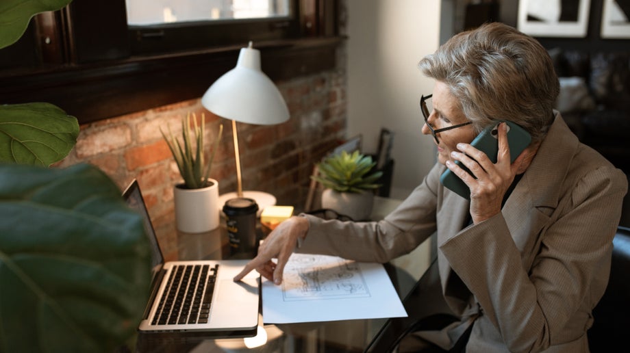 An individual is talking to someone on their mobile phone while they sit in front of their desk and use their laptop at the same time.