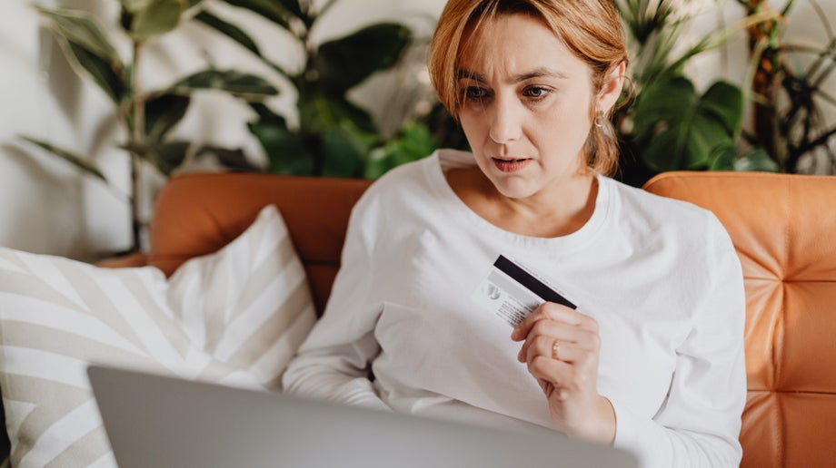 An individual is sitting down on a couch with a laptop placed on top of their legs. They are viewing the screen while they are holding a card in their hand.