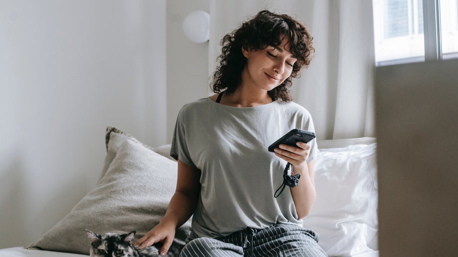 An individual is sitting on their bed while holding and viewing their mobile screen with one hand and petting their cat with the other hand.