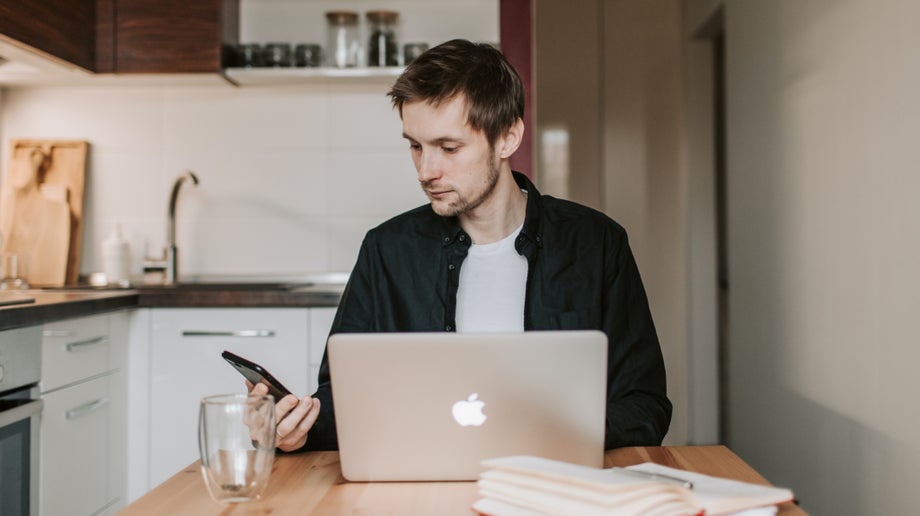 An individual is sitting at their kitchen table while using their mobile phone. Their laptop is in front of them.