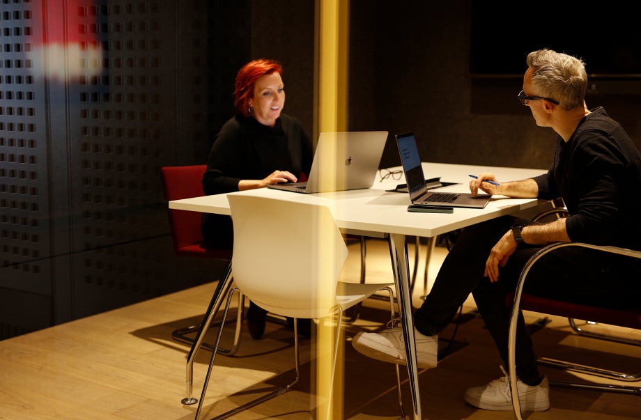 Two individuals are in a meeting room, sharing a desk. They are sitting across from one another, talking and working on their laptops which are in front of them.