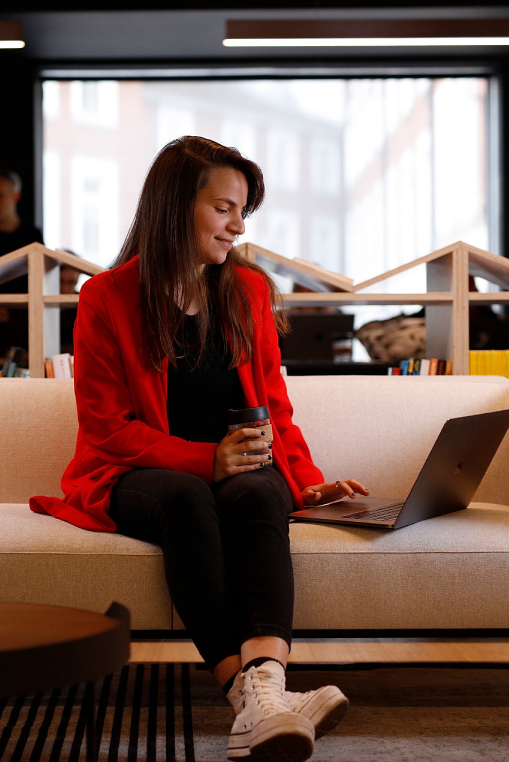 An individual in a bright red jacket is sitting down on a modern couch while they work on their laptop with one hand and hold a coffee cup in the other.