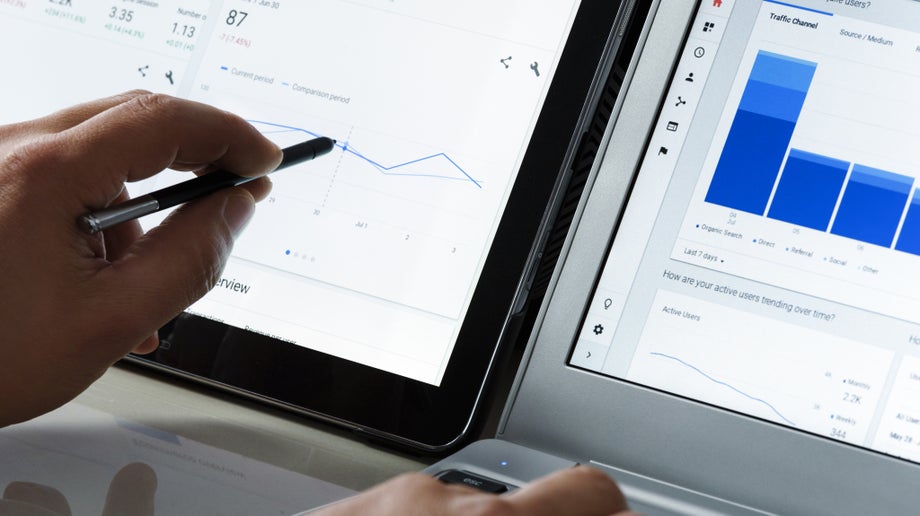 A close up of a hand interacting with his tablet on the left and his laptop on the right. The two devices are placed side-by-side and statistics can be seen on both screens.