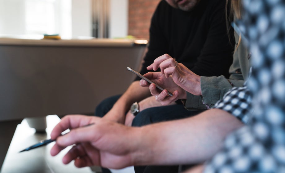 Three people sit in a line, one pointing to their phone, another using a pen to point at something on the table out of view. 