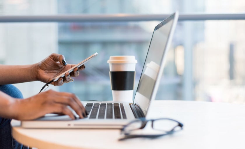 A person types on their laptop, reading from a mobile phone 