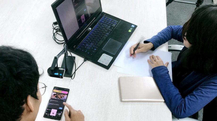 User testing is taking place. An individual is testing a mobile device while the tester monitors the test on their laptop.