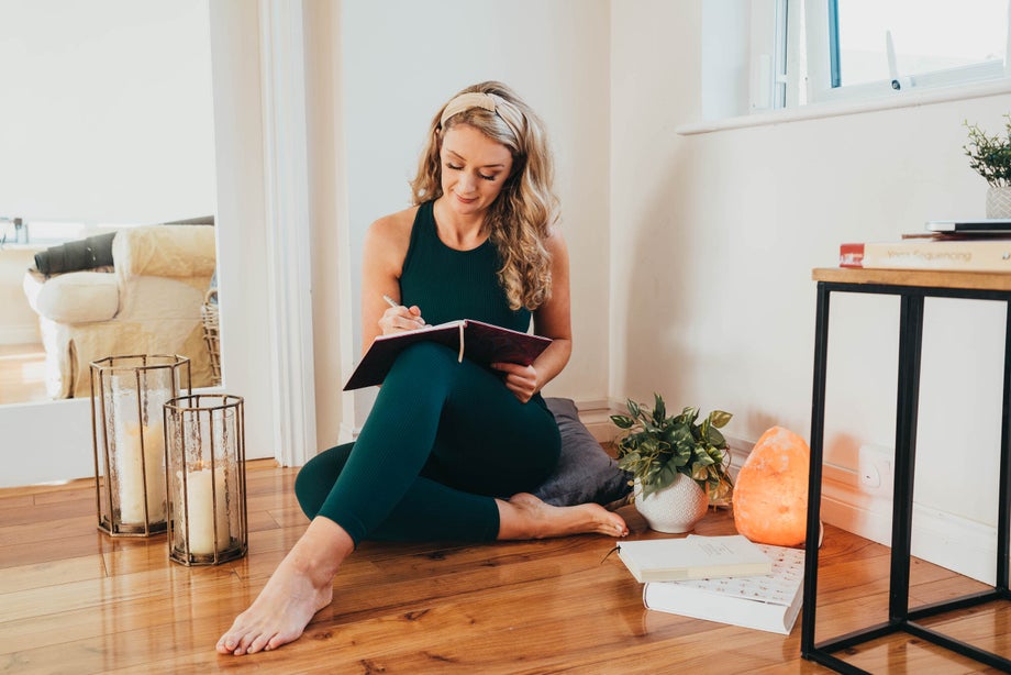 A woman sits barefoot on a wooden floor writing in a notebook