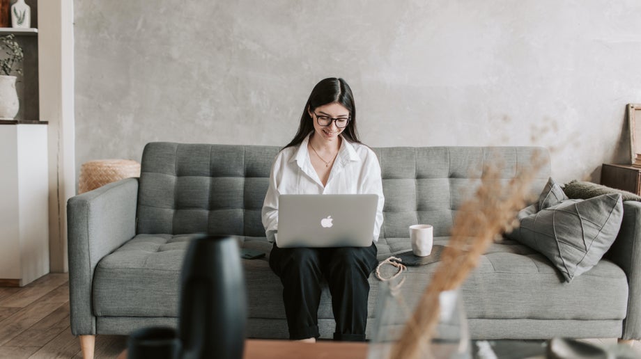 An individual is sitting in the middle of their couch while they use their laptop which is resting on their legs.