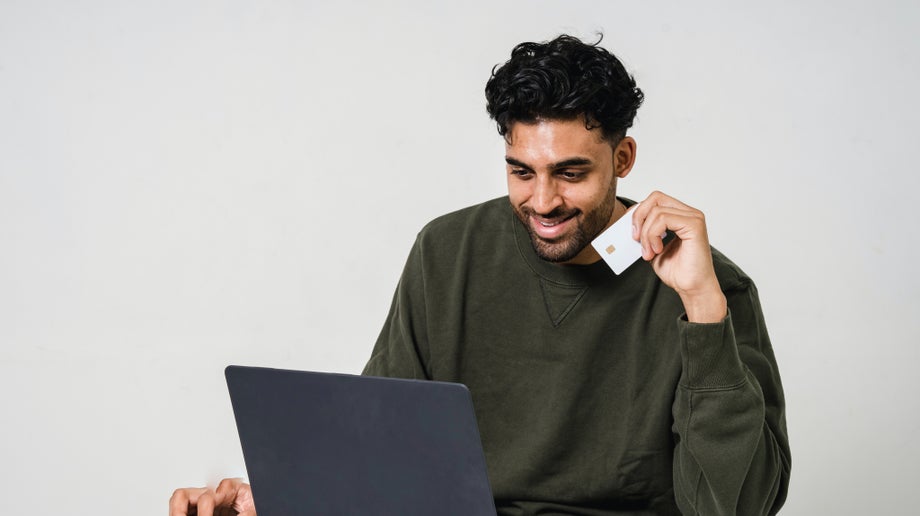 An individual is sitting at a table looking down at their laptop while one hand is using the laptop’s mouse and the other is holding a credit card.